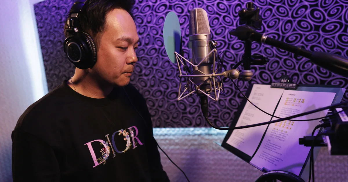 Voice actor on headphones reading lines from a script stand at a large-diaphragm condenser microphone in the MIX I booth